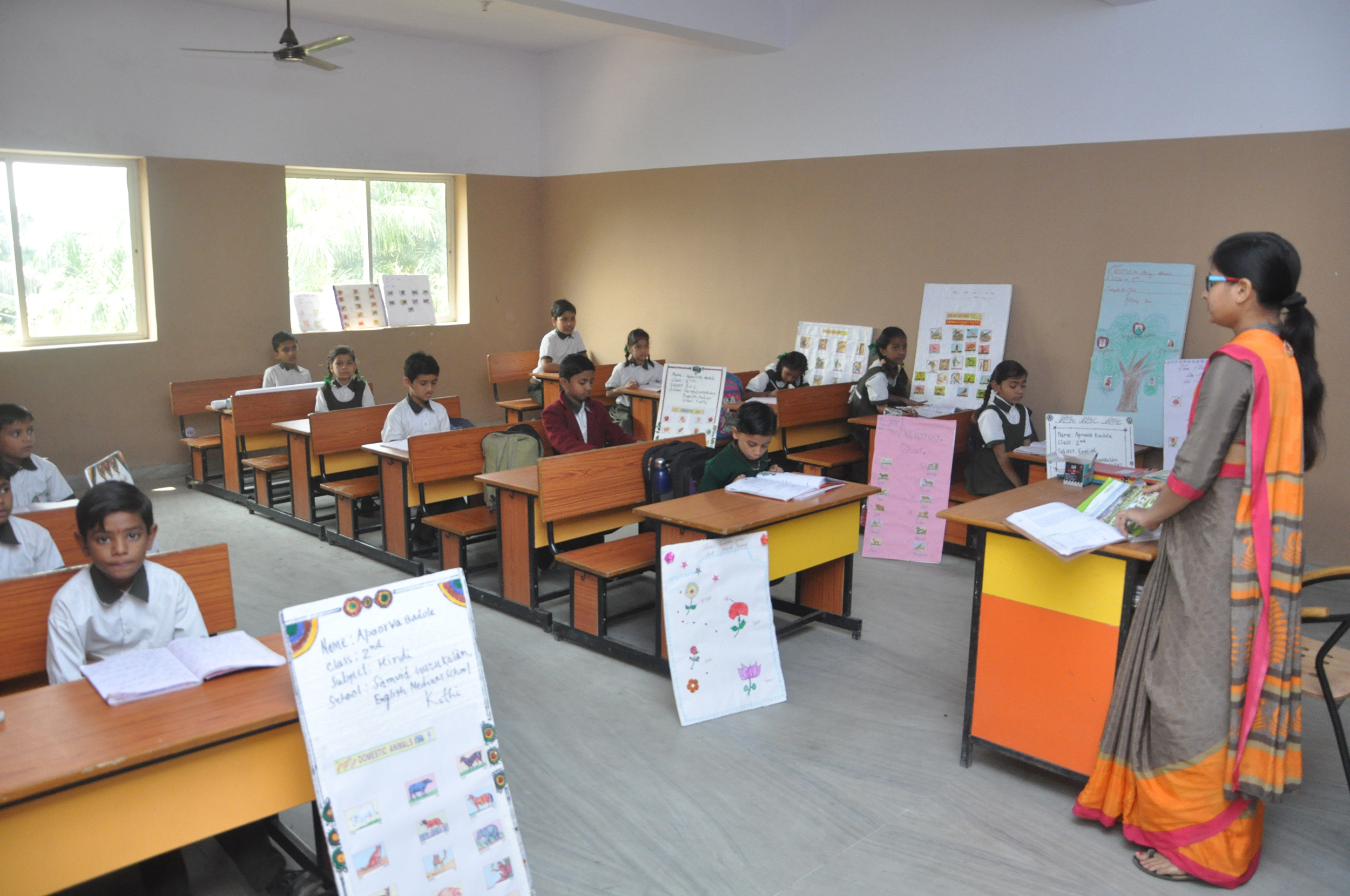 children sitting at desks with books open while their teacher speaks to them