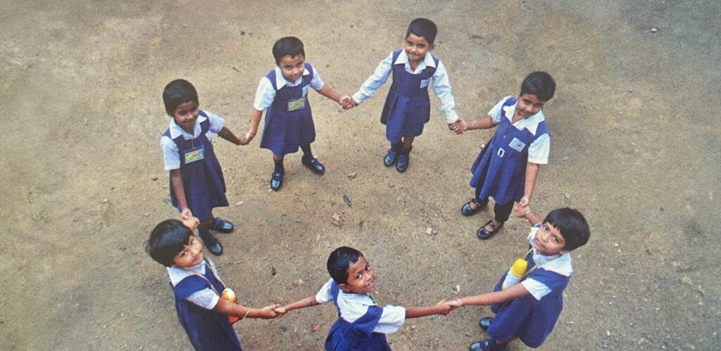 Schoolchildren holding hands in a circle looking up at the camera in rural India