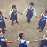 Schoolchildren holding hands in a circle looking up at the camera in rural India