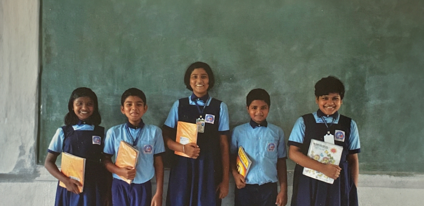 children holding school books in a line against a chalkboard smiling at the camera