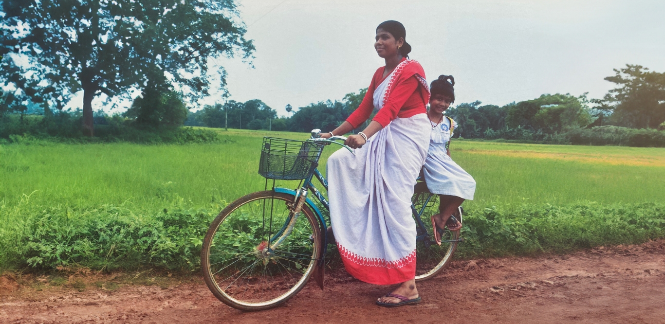 mother riding a bicycle on a dirt road with a child sitting on the back in rural India